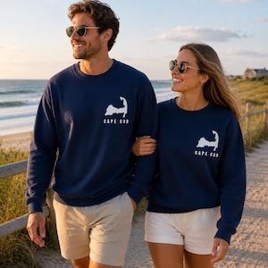 May include: Two people wearing navy blue sweatshirts with a white Cape Cod outline and text. They are walking on a sandy path near the ocean. The sweatshirts have long sleeves and a crew neck.