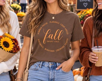 May include: A brown short-sleeve t-shirt featuring the word fall in large orange script, with the text sweet fall below it, and a circular border of words including pumpkin patch, flannels, crisp apples, crunchy leaves, bonfires, and football.