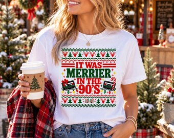 May include: A white short-sleeved t-shirt featuring a festive graphic with the text IT WAS MERRIER IN THE 90'S, surrounded by red and green patterns, cassette tapes, and holiday lights, designed in a classic holiday sweater style.