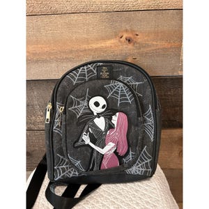 Op de afbeelding: Zwarte denim rugzak met Jack Skellington en Sally uit The Nightmare Before Christmas. De rugzak heeft een ritssluiting, verstelbare bandjes en een webpatroon. De Disney- en Loungefly-logo's zijn zichtbaar.