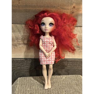 May include: A doll with bright red, voluminous hair and large, expressive eyes. The doll is wearing a pink and white checkered dress. The doll is standing on a textured surface, with a wooden background.