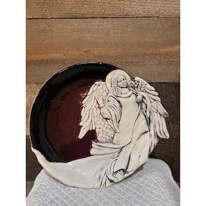 May include: A round ceramic plate featuring a raised, sculpted white angel figure with detailed wings and flowing robes. The background of the plate is a deep, glossy maroon color, while the angel is finished in a creamy white glaze with dark accents.