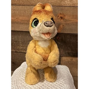 Op de afbeelding: Een vintage pluche speelgoed in de vorm van een cartoon dier, mogelijk een hond of vos. Het speelgoed heeft een geel en wit kleurenschema met grote, expressieve ogen en een glimlachende uitdrukking. Het staat op een wit oppervlak tegen een houten achtergrond.