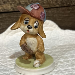 May include: A ceramic figurine of a brown puppy wearing a blue and brown baseball cap with stitched patches. The puppy has large eyes, a baseball glove on one paw, and stands on a round green base next to a small white baseball.