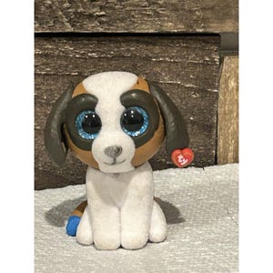 May include: A small, plush dog figurine with large, sparkly blue eyes. The dog has brown and white fur, with dark brown ears and a red heart-shaped tag. The figurine is sitting, with a blue accent on one paw.