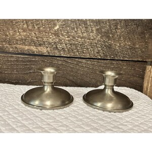 Puede incluir: Dos candeleros de metal plateado. Cada uno tiene una base redonda y un tallo cilíndrico corto con una copa poco profunda para sostener una vela. Los candeleros se exhiben sobre una superficie blanca texturizada contra un fondo de madera.