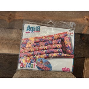 Può includere: Un materassino gonfiabile Tropical Air Mat di Aqua Leisure confezionato. Il materassino presenta un colorato motivo floreale nei toni del rosa, blu, giallo e arancione. La confezione include il testo "Aqua Leisure" e "Tropical Air Mat".