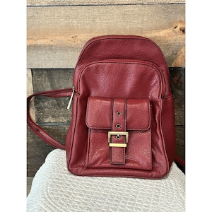 Puede incluir: Una mochila roja con un acabado texturizado. La mochila tiene un bolsillo frontal con una solapa asegurada por una hebilla dorada. Tiene un cierre de cremallera superior y correas ajustables.