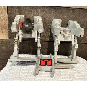 May include: Three light grey toy robots with articulated legs and a central body. One robot has a black and red component, and another has a red gear. The robots are on a white surface with a wooden background.