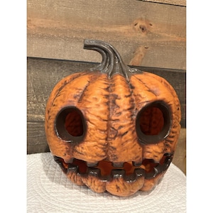 May include: A carved orange pumpkin with a dark gray stem and dark brown eye and mouth openings. The pumpkin has a textured surface and is likely used as a Halloween decoration.
