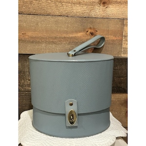 Könnte beinhalten: Eine hellblaue Hutschachtel im Vintage-Stil mit strukturierter Oberfläche. Die runde Box hat einen Griff und einen goldfarbenen Verschluss. Die Box steht auf einer weißen, spitzenartigen Oberfläche mit Holzgrund.