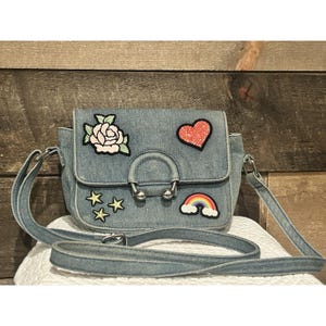 May include: A light blue denim crossbody bag with a flap closure and a long adjustable strap. The bag is decorated with embroidered patches of a rose, a glitter heart, stars, and a rainbow. The bag has a silver-tone metal clasp.