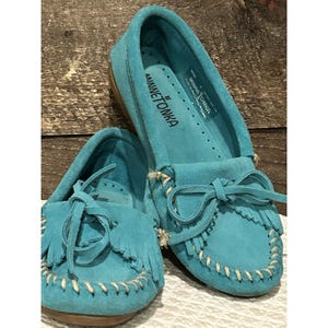 May include: Turquoise suede moccasins with fringe and bow details. The shoes have the brand name "MINNETONKA" printed on the insole. The soles are a light brown color, and the stitching is a cream color.