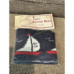 May include: A navy blue knitted band with a sailboat design. The sailboat has a white sail, red accents, and is set against a navy blue background. The band is packaged with a label that reads "Lyle's Knitted Band."