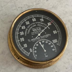 May include: A round brass-cased Abbeon certified hygrometer and temperature indicator with a black face. The dial displays relative humidity from 0 to 100 percent and temperature in Fahrenheit from 0 to 180 degrees, with white text and needles.