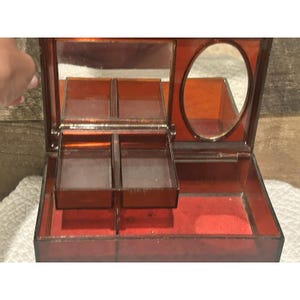 Op de afbeelding: Een open, vintage sieradendoosje met een transparante, roodbruine buitenkant. De doos heeft een rechthoekige vorm met een scharnierend deksel en meerdere compartimenten, waaronder een kleine ovale spiegel en een rode fluwelen binnenkant.