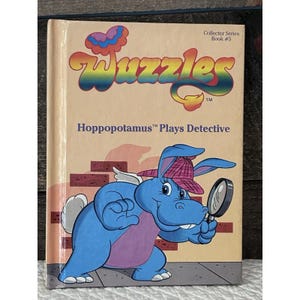 Könnte beinhalten: Ein Vintage-Kinderbuch mit dem Titel "Wuzzles Hoppopotamus Plays Detective". Das Cover zeigt einen blauen Nilpferd-Charakter mit Detektivhut und Lupe. Das Buch hat einen beigefarbenen Einband mit farbenfrohem Text und Illustrationen.