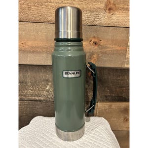 May include: A green Stanley thermos with a silver lid and base. The thermos has a handle and the Stanley logo is visible on the front. The thermos is sitting on a white cloth in front of a wooden background.