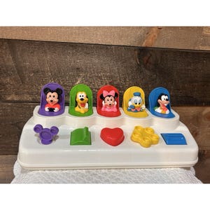 以下が含まれることがあります： ミッキーマウス、プルート、ミニーマウス、ドナルドダック、グーフィーの5人のディズニーキャラクターが特徴の白いプラスチック製ポップアップおもちゃ。各キャラクターは色付きのスロットに収まっており、その下には紫色のミッキーアイコン、緑色のくさび形、赤色のハート、黄色の花、青色の長方形などの対応する形状が配置されています。