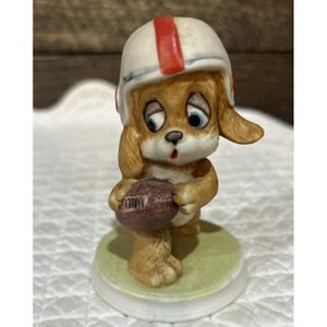 May include: A ceramic figurine of a brown puppy with large blue eyes wearing a white football helmet with a red stripe. The puppy is holding a textured brown football in its paws and stands on a round, light green base.