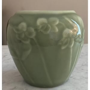 May include: A sage green ceramic vase with a rounded, bulbous shape and a wide opening. The exterior surface features a raised, embossed floral pattern of delicate flowers and stems that wrap around the body of the vessel in a monochromatic finish.
