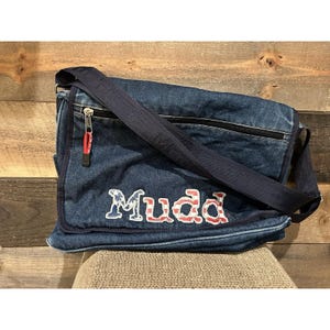 May include: A blue denim messenger bag with a long adjustable strap. The bag features the word "Mudd" in a patriotic font with red, white, and blue accents. It has a zipper closure and a red zipper pull.