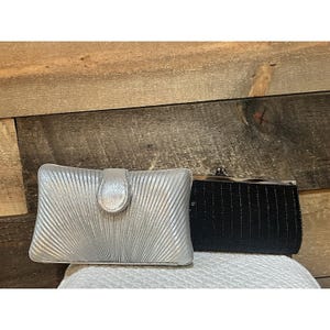 May include: A silver clutch with a pleated fan-like texture and a central tab closure, positioned next to a black rectangular clutch featuring a fine ribbed surface and a metallic kiss-lock frame closure at the top edge.