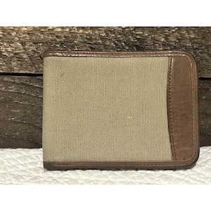 May include: Tan canvas wallet with brown leather accents. The wallet has a rectangular shape with a bi-fold design. The leather is on the edges and a vertical panel. The background is a wooden surface.