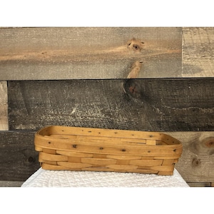 May include: A rectangular, light brown woven basket with rounded corners. The basket is made of thin strips of wood, held together with small metal fasteners. The basket is empty and sits on a white, textured surface.