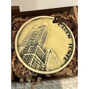 May include: A round, cream-colored ornament featuring a detailed illustration of the Carew Tower building. The building is depicted in black and white, with the text "Carew Tower" in a circular pattern around the edge. The ornament has a brown ribbon for hanging.