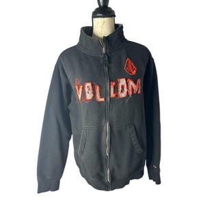 May include: Black zip-up hoodie with the word "VOLCOM" in red and white stitched lettering across the chest. A red and white logo is on the upper right chest. The hoodie has a high collar and two front pockets.