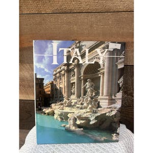 May include: A hardcover book titled ITALY featuring a photographic cover of the Trevi Fountain in Rome. The image displays the ornate stone architecture, classical sculptures, and turquoise water of the fountain under a bright blue sky.