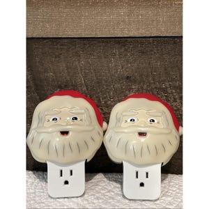 Peut inclure: Deux veilleuses Père Noël. Chaque veilleuse présente un visage blanc avec un bonnet rouge et une barbe blanche. Les veilleuses se branchent directement dans une prise électrique standard.