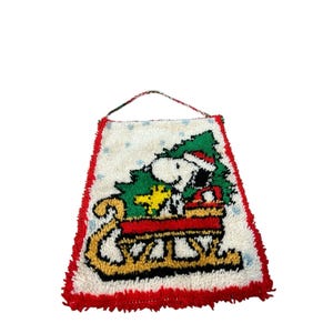 Op de afbeelding: Een rechthoekige wandhanger in knooptechniek met het witte Snoopy-personage in een rode kerstmuts, zittend in een gele slee met een groene kerstboom en een geel Woodstock-figuurtje, tegen een witte achtergrond met blauwe sneeuwvlokken.