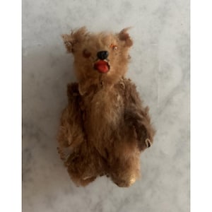 May include: A small, vintage-style teddy bear crafted from brown, textured fur. The bear features a black stitched nose, a bright red mouth, and small, dark, inset eyes. Its limbs are positioned at its sides, and the fur appears worn and matted.