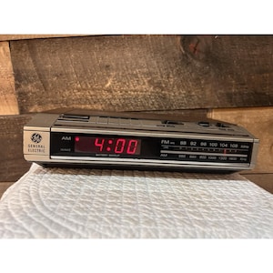 Puede incluir: Un radio reloj despertador digital General Electric con carcasa plateada y negra. La pantalla frontal muestra un reloj digital rojo brillante que marca las 4:00, un sintonizador de radio AM y FM, y el texto Battery Backup impreso debajo.