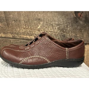 May include: Brown leather slip-on shoe with decorative stitching. The shoe has a textured surface and a black sole. The stitching is a contrasting off-white color. The shoe is designed for comfort and casual wear.
