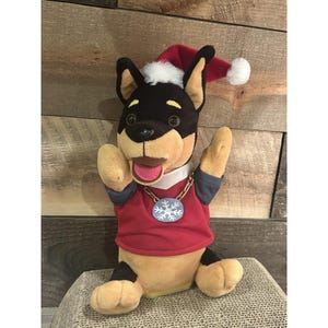 Puede incluir: Un juguete de peluche con forma de perro que lleva un gorro de Navidad rojo con un pompón blanco y una camisa roja. El perro tiene pelo negro y canela, un collar de cadena dorada con un colgante de copo de nieve.