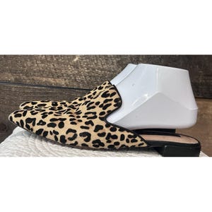 May include: A pair of slip-on mules with a leopard print pattern. The shoes have a pointed toe and a black sole. The upper part of the shoe is tan with black spots. The shoes are displayed on a white shoe form.