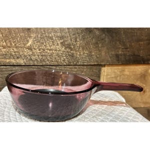 May include: A translucent purple glass saucepan with a round bowl and a single integrated handle extending from the side. The glass features a smooth, polished finish and a deep violet hue that allows light to pass through the material.