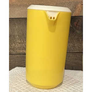 以下が含まれることがあります： 白い蓋が付いた黄色のプラスチックピッチャー。ピッチャーには注ぎ口と丸みを帯びたベースがあります。背景は木製の表面です。ピッチャーはおそらく飲み物を提供するのに使用されます。