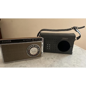 May include: A vintage Zenith Royal 510 transistor radio with a brown front panel, silver trim, and a circular AM/FM dial. Beside it sits a black protective carrying case with a perforated speaker grill and an adjustable leather shoulder strap.