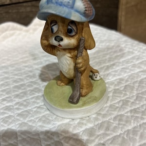 Puede incluir: Una figura de cerámica de un cachorro marrón con ojos grandes y tristes que lleva una gorra de cuadros azules. El cachorro está sobre una base redonda de color verde claro, sosteniendo un palo de golf marrón. Una pequeña pelota de golf blanca con puntos negros se encuentra cerca de sus patas traseras.