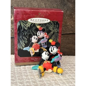 Può includere: Una scatola di ornamenti Keepsake con statuette di Topolino e Minnie. Le statuette indossano pattini e abiti invernali. La scatola è rossa con scritte dorate e un'immagine delle statuette. Il testo "New Pair of Skates" è visibile.