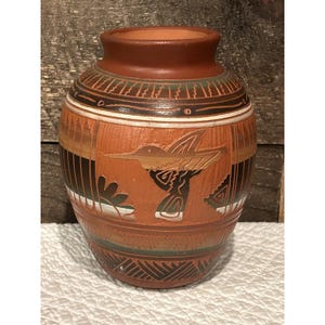 May include: A terracotta-colored ceramic vase with a hummingbird design. The vase features a cylindrical shape with a narrow neck and a wide base. The hummingbird is carved into the surface, along with other geometric patterns and designs.