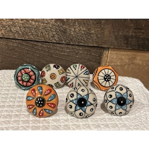 May include: A collection of eight decorative ceramic cabinet knobs. The knobs feature various colorful patterns, including floral, geometric, and abstract designs. Colors include orange, blue, green, and white. The knobs are round and have a glossy finish.