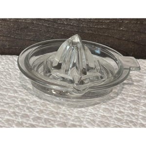 May include: A clear glass citrus juicer with a ridged reamer and a circular base. The juicer has a spout for easy pouring. The design is simple and functional, perfect for extracting fresh juice. The juicer is made of clear glass.
