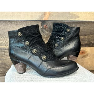 May include: A pair of black leather ankle boots with decorative floral accents and a textured suede panel. The boots have a low heel and are displayed on a white surface.