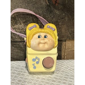 May include: A vintage yellow toy radio with a Cabbage Patch Kids design. The radio has a light pink handle, a round dial, and musical note graphics. The face of the radio is a Cabbage Patch Kids doll head with blonde hair and blue eyes.