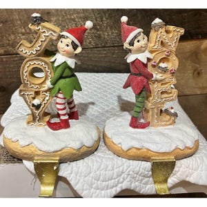 May include: Two Christmas stocking holders featuring elves standing on snow-covered bases. One elf wears a green outfit and holds the letters JOY, while the other wears a red outfit and holds the letters NOEL. Both elves wear red and white hats.